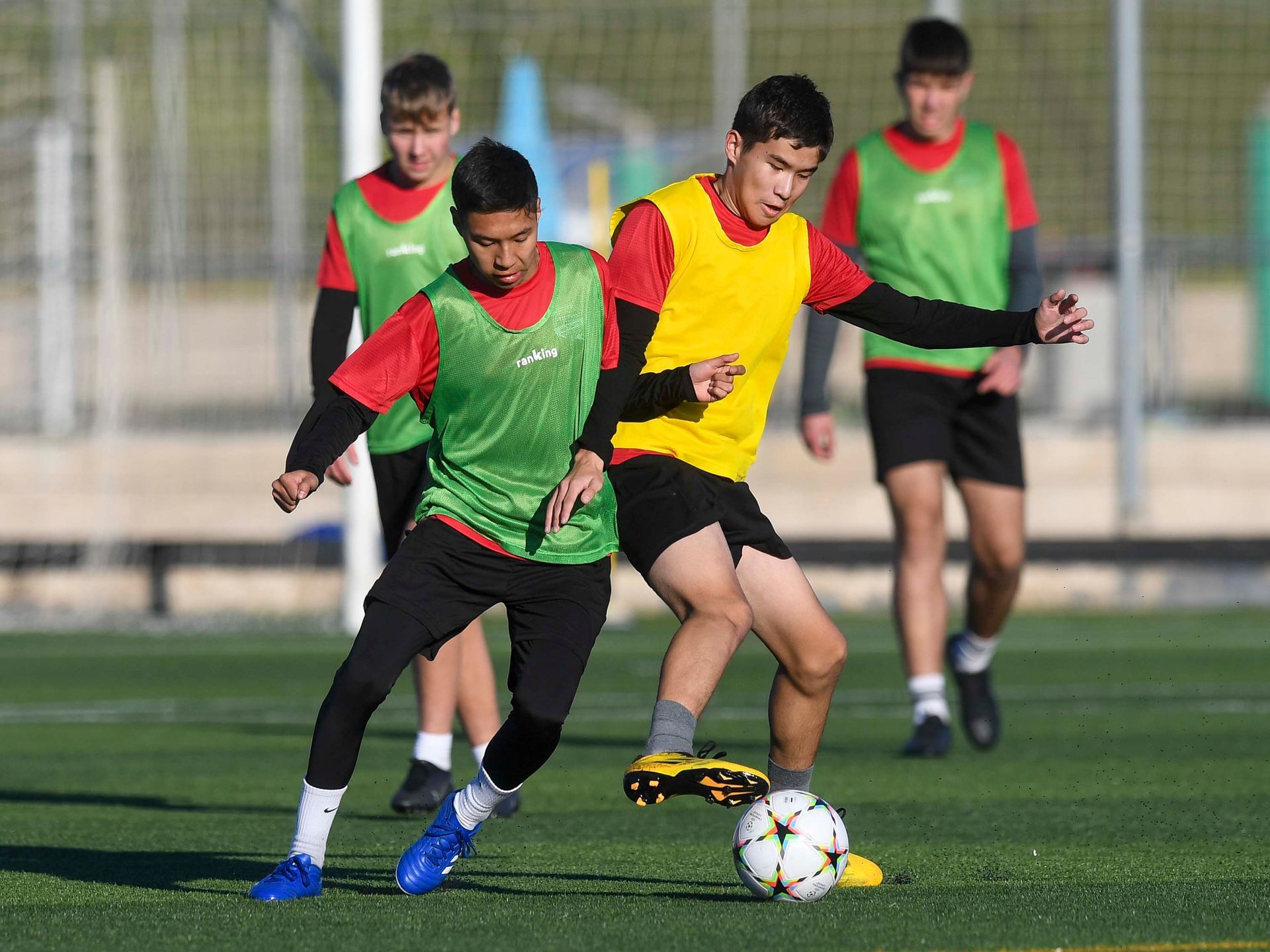 Los jugadores de fútbol con chalecos verdes y amarillos practican en un campo, mientras uno de ellos dribla el balón.
