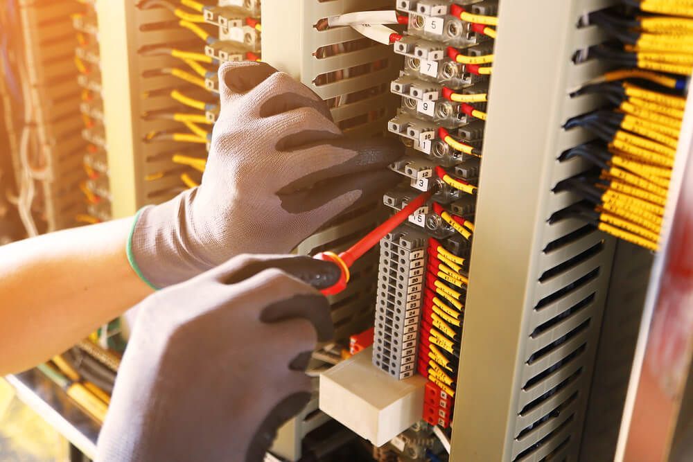 Des mains gantées utilisent une pince coupante rouge sur un panneau de commande électrique dont les fils et les bornes sont étiquetés.