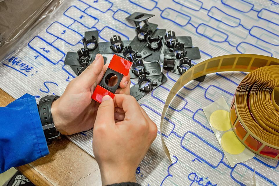Des mains assemblent un petit composant électronique rouge sur un établi, parmi des composants noirs éparpillés et une bobine de fil.