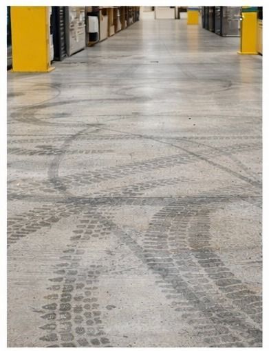 Huellas de neumáticos en el suelo de un almacén de hormigón, filas de estantes en el fondo.