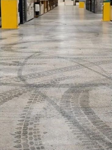 Huellas de neumáticos en el suelo de un almacén de hormigón, filas de estantes en el fondo.