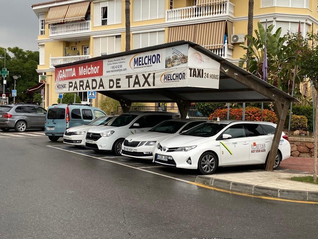Parada de taxis con varios coches aparcados bajo una marquesina; edificio al fondo.
