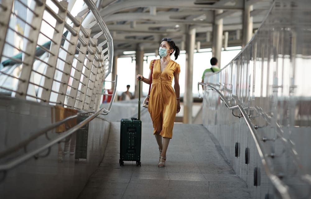 Mujer con vestido amarillo y mascarilla tirando de una maleta en una pasarela moderna.