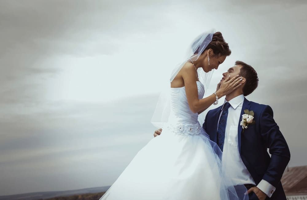 Los novios se miran fijamente, la novia le toca la cara. Fondo de cielo nublado.