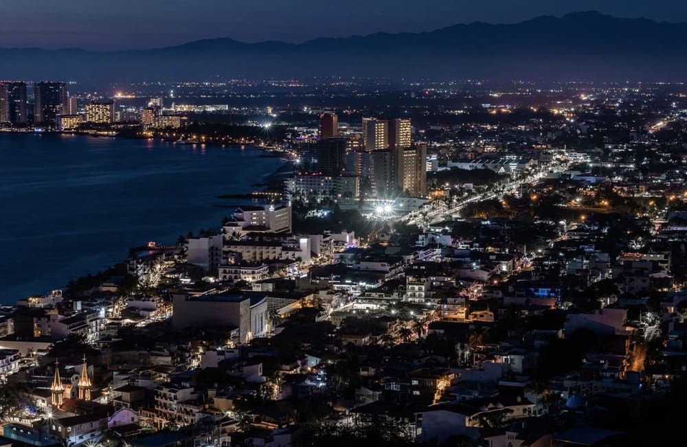 Vista nocturna de una ciudad costera con luces centelleantes a lo largo de la costa y montañas al fondo.