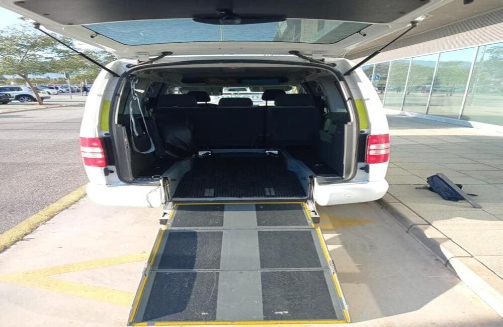 Furgoneta blanca con puerta trasera abierta y rampa para silla de ruedas desplegada en la acera.