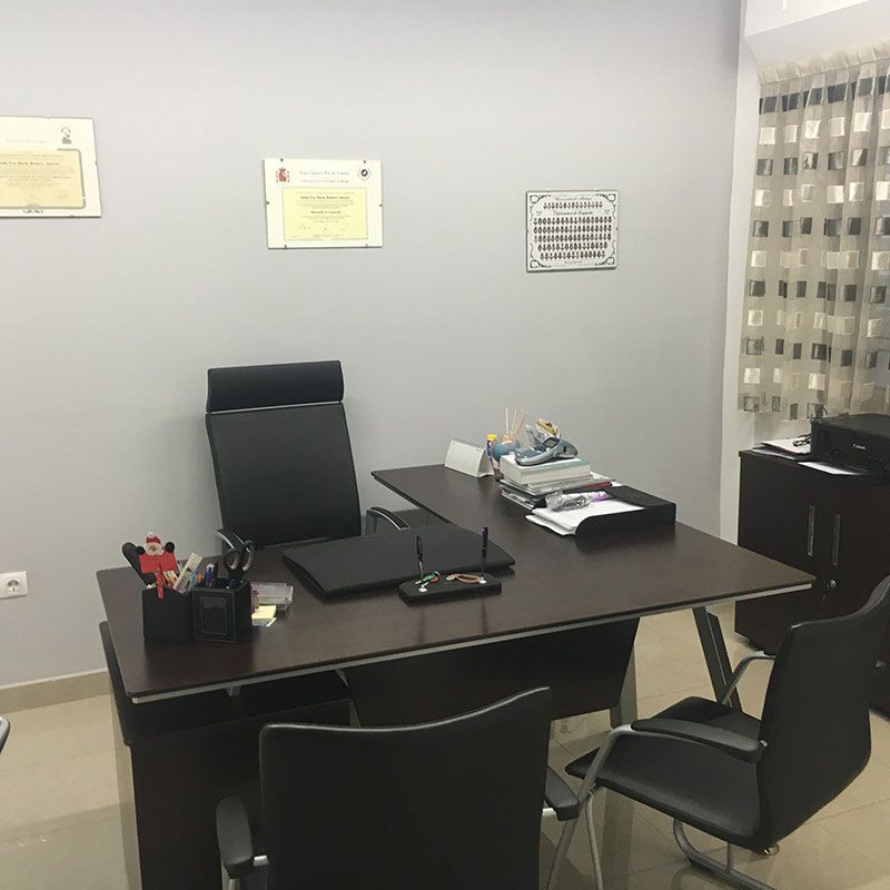 Oficina vacía con escritorio oscuro, dos sillas y documentos en una pared gris claro.