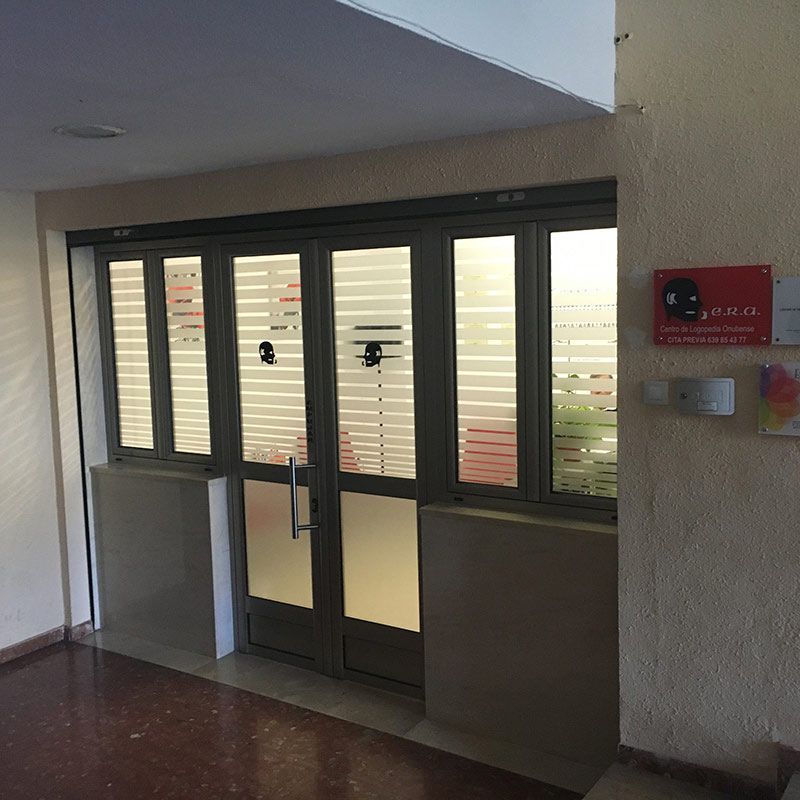 Entrada cubierta al edificio con puertas dobles de cristal oscuro, ventanas laterales y un letrero rojo en la pared.