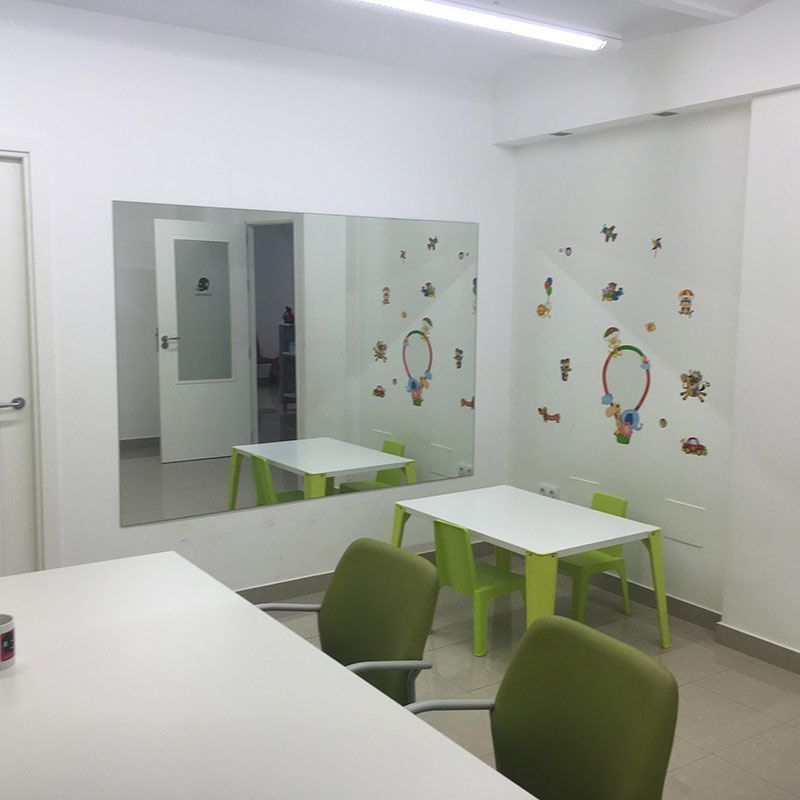 Aula luminosa con sillas verdes, mesas blancas, espejo de pared y vinilos decorativos de mariposas.
