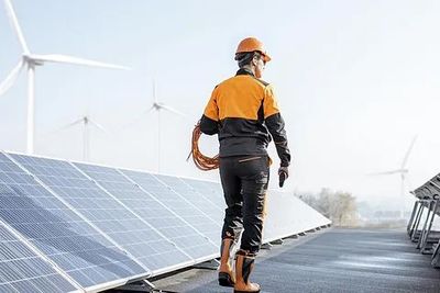 Un trabajador con equipo de seguridad naranja inspecciona paneles solares en un tejado, con aerogeneradores al fondo.
