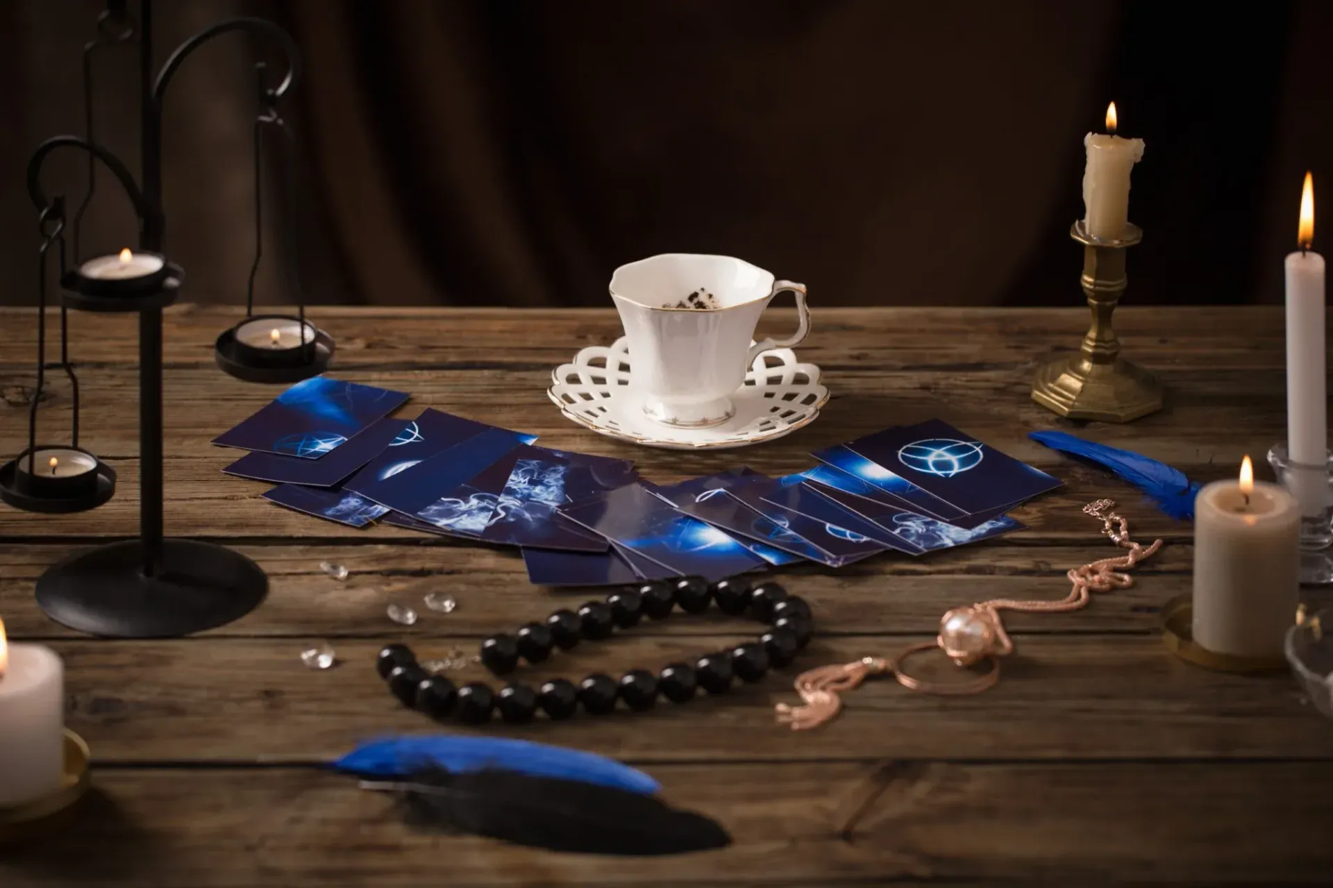 Escena de lectura de cartas del tarot: cartas, taza de té, velas, cuentas oscuras, plumas sobre mesa de madera.