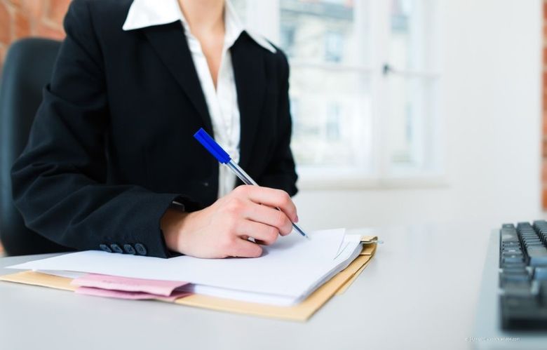 Eine Person in einem schwarzen Blazer schreibt an einem Schreibtisch in ein Notizbuch; eine Tastatur befindet sich in der Nähe.