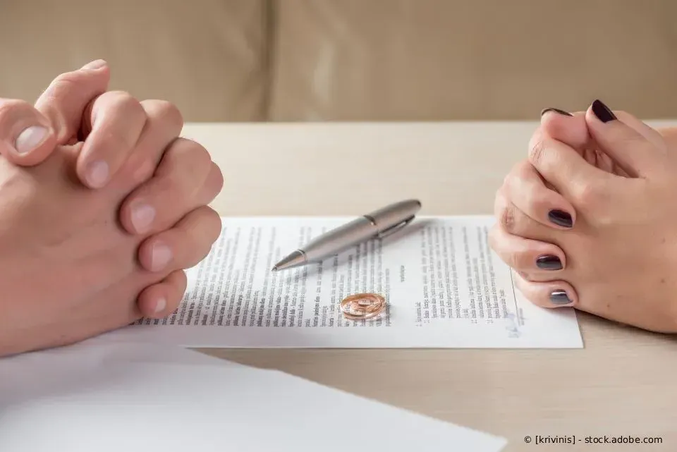 Zwei Personen sitzen mit verschränkten Händen an einem Tisch; neben ihnen liegen ein Stift, ein Dokument