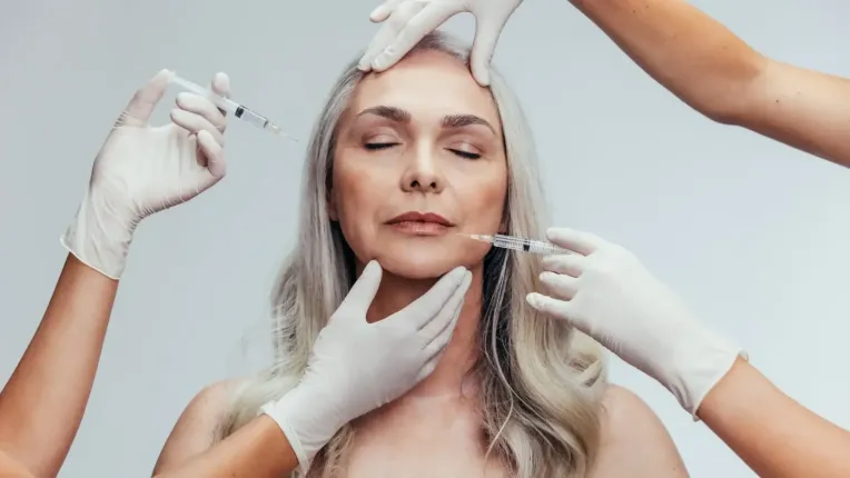 Mujer recibiendo inyecciones cosméticas rodeada de manos enguantadas, en el interior.
