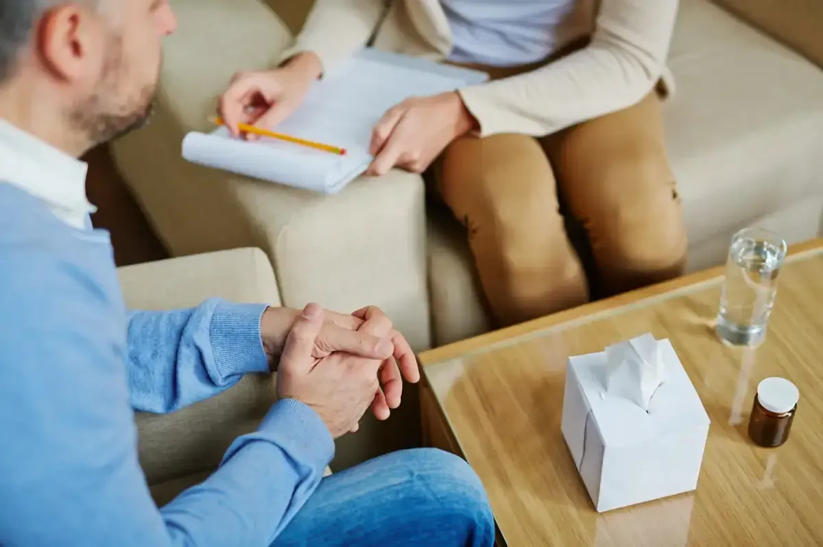 Hombre en sesión de terapia, con las manos entrelazadas, frente al terapeuta con un bloc de notas.