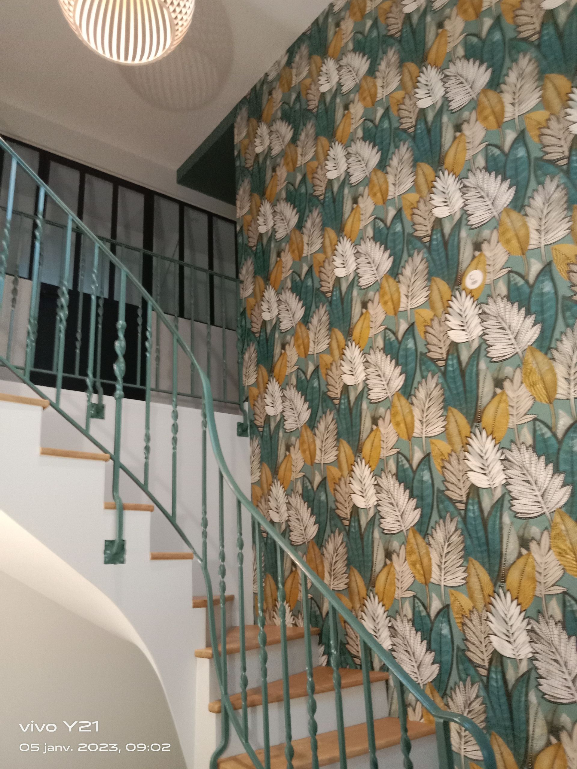 Escalier avec rampe bleu sarcelle, papier peint fleuri dans un hall avec un motif de feuilles blanches et jaunes.