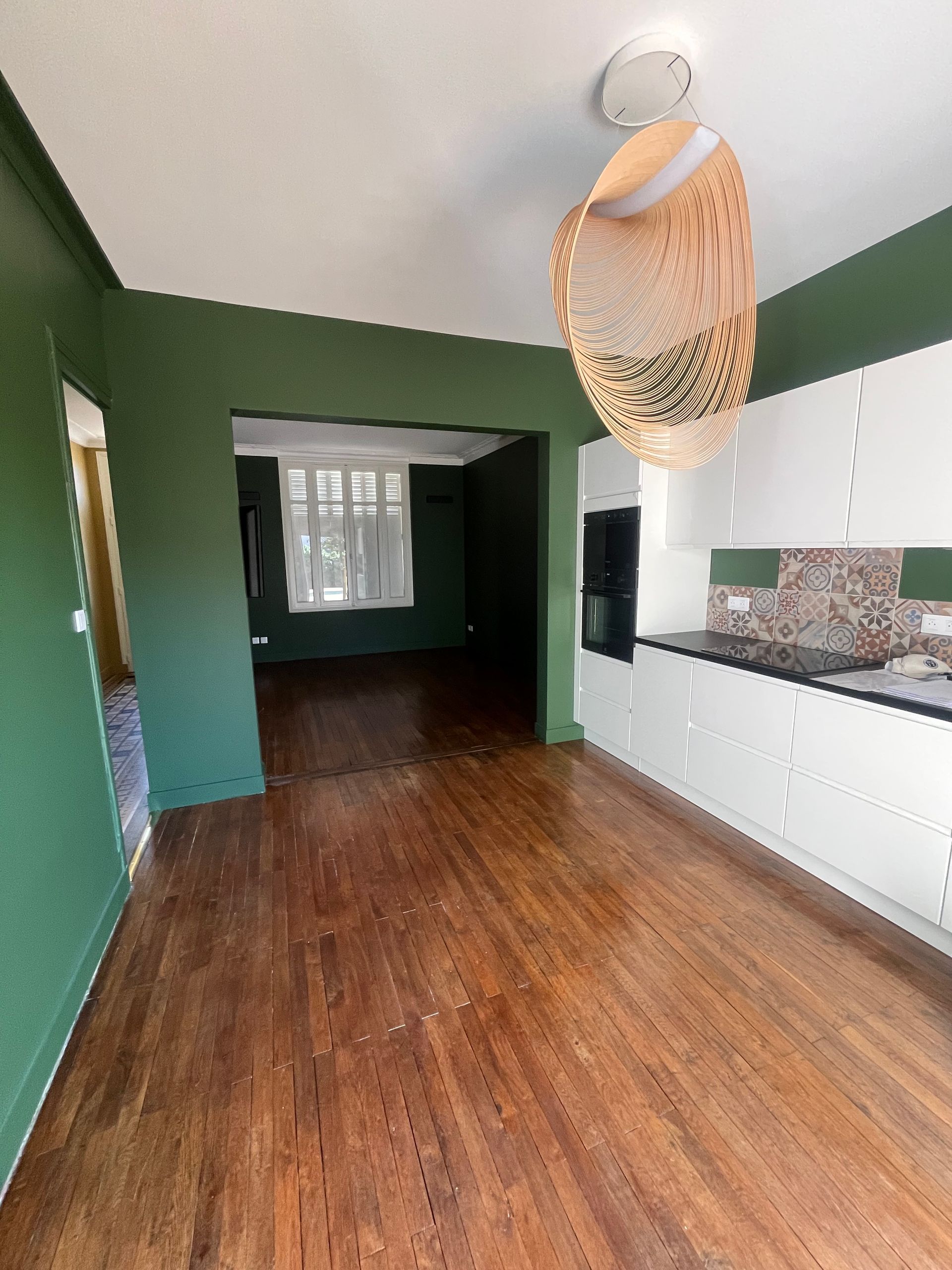 Pièce vide aux murs verts, avec un parquet en bois et des placards blancs sous un luminaire suspendu en fibres tressées.