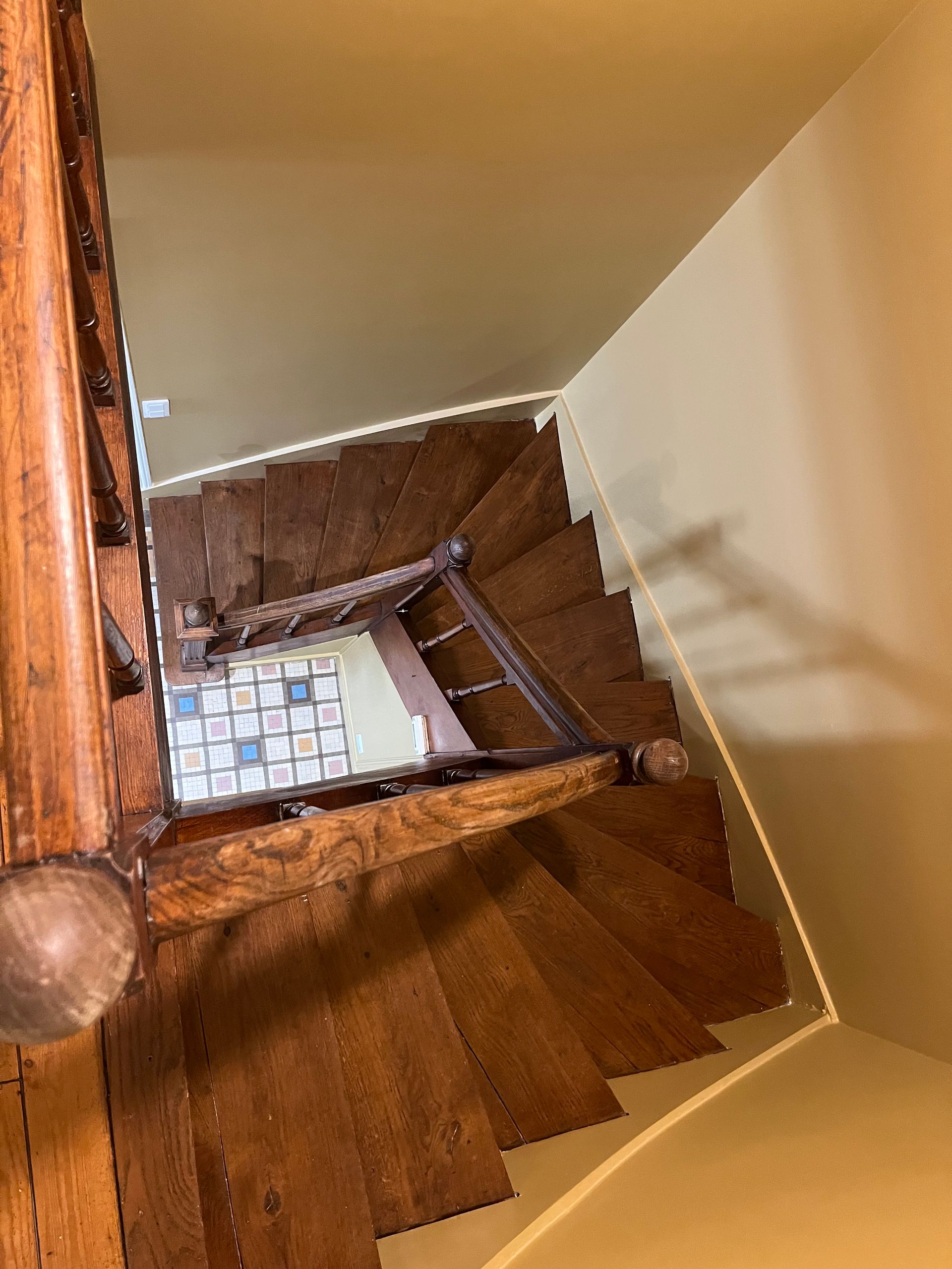 Vue plongeante sur un escalier en bois avec des murs beiges et un palier carrelé en contrebas.