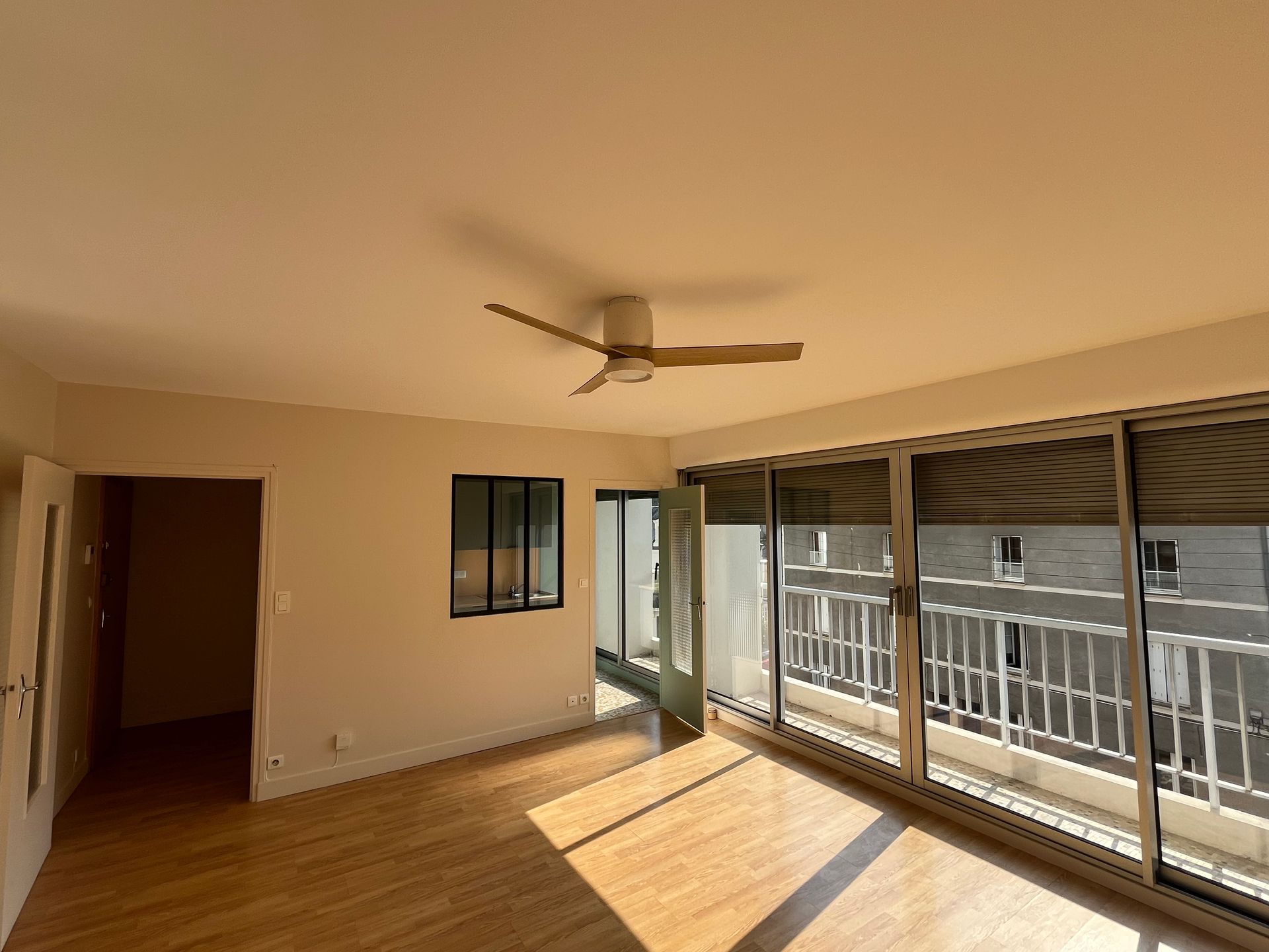Salon vide avec parquet, ventilateur de plafond et portes-fenêtres coulissantes donnant sur un balcon.