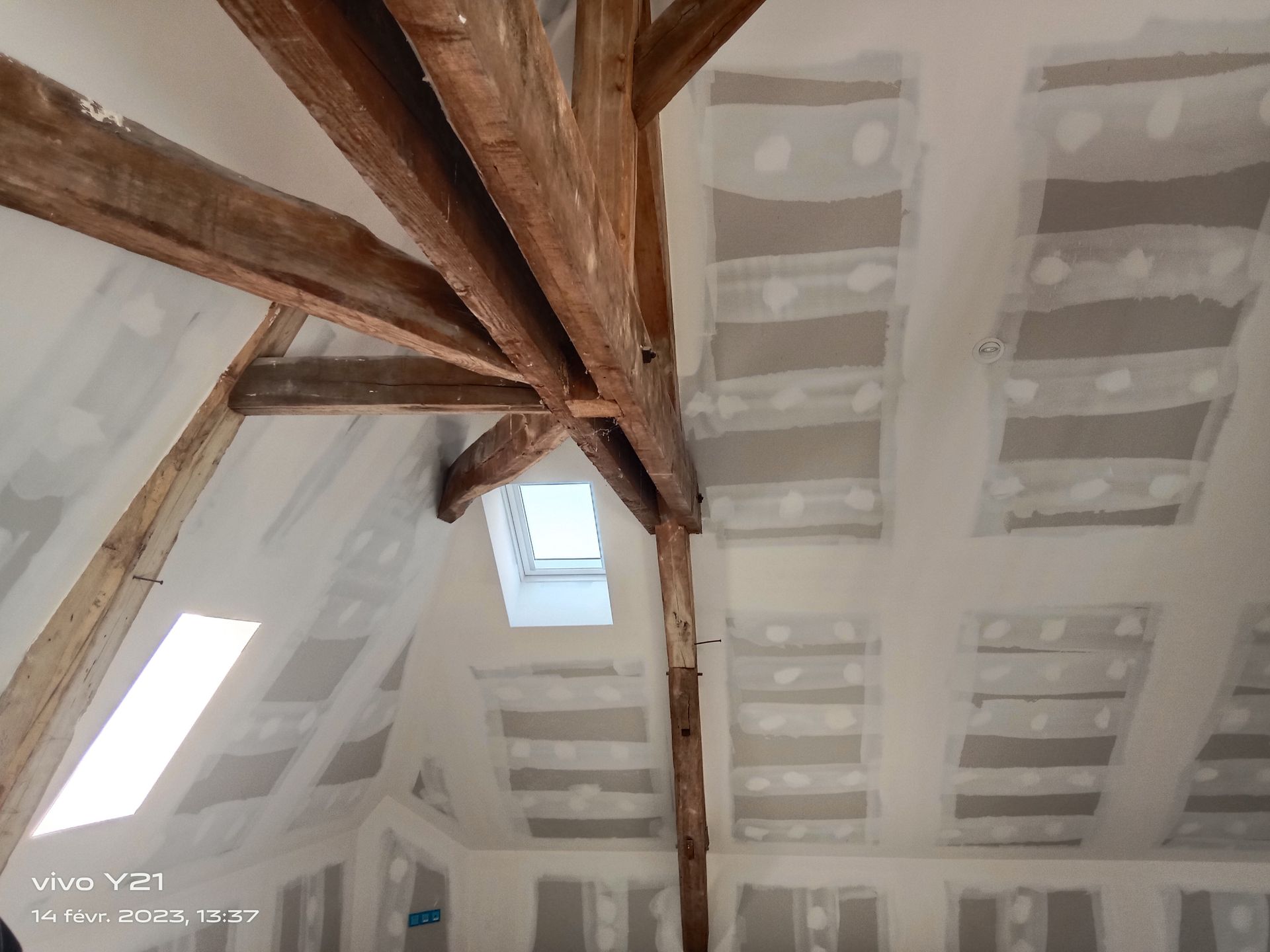 Intérieur d'un grenier rénové avec poutres en bois apparentes, cloisons sèches et puits de lumière.