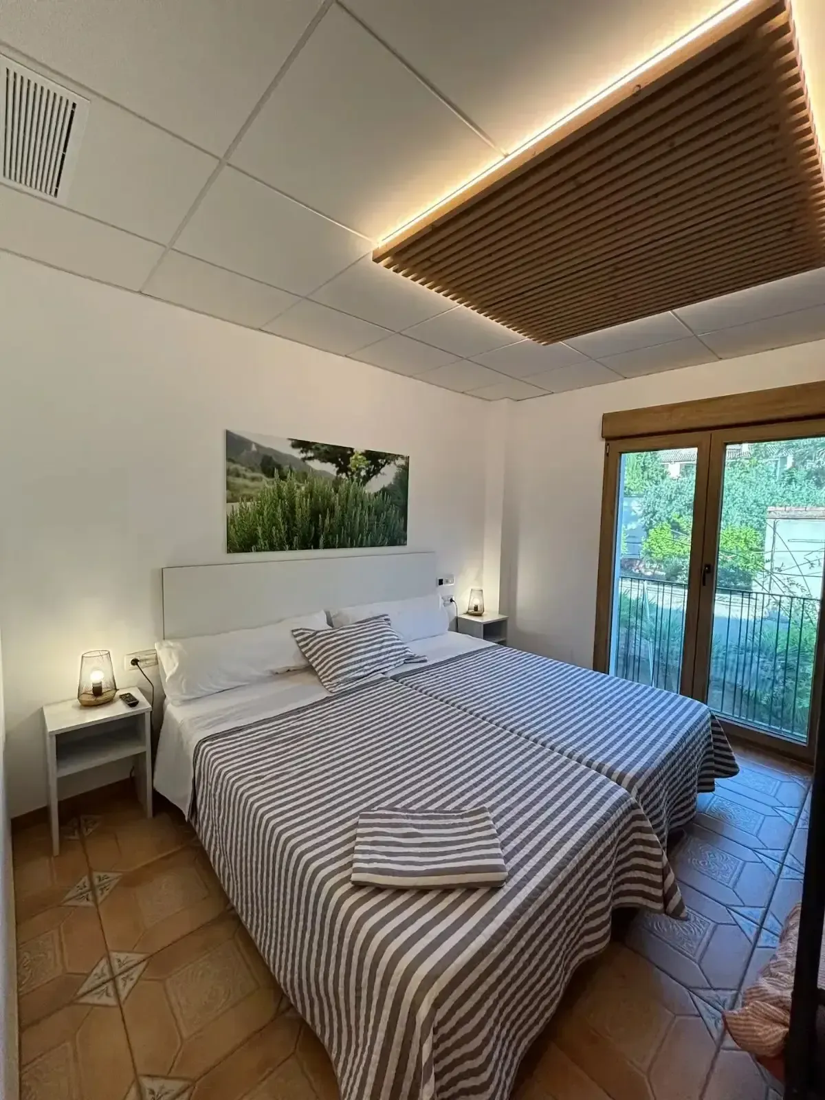 Dormitorio con dos camas individuales, colchas de rayas, paredes blancas, panel de techo de madera y vista a un jardín.