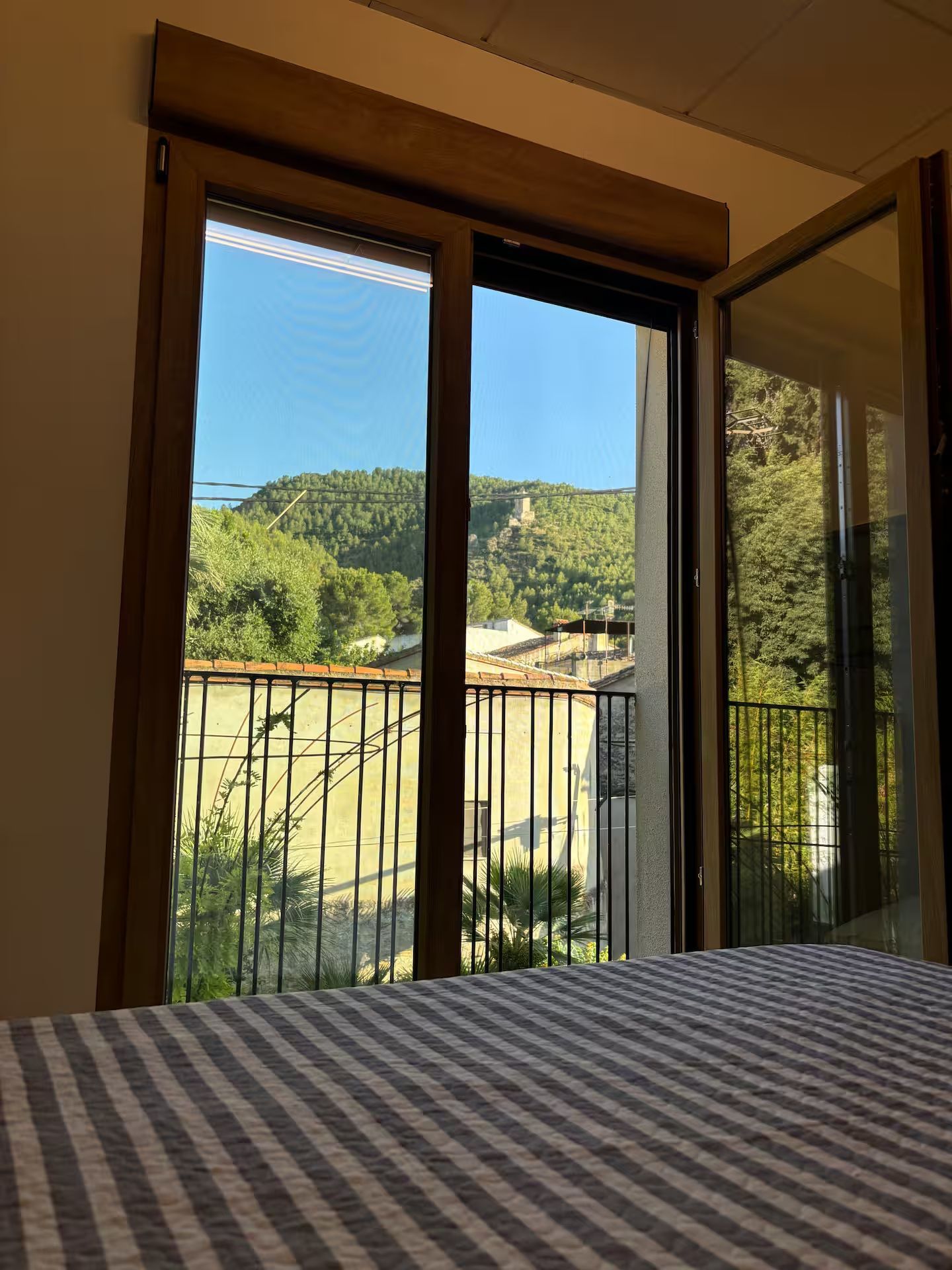 Ventana del dormitorio abierta, con vista a un paisaje verde con montaña, cielo azul y un balcón de hierro forjado.