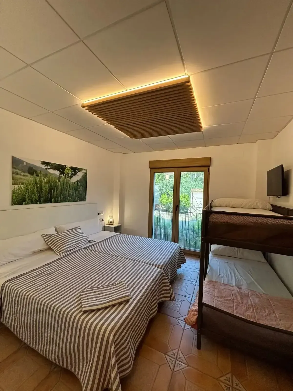 Dormitorio con dos camas individuales, una litera y una puerta a un patio; techo de madera clara; arte paisajístico.