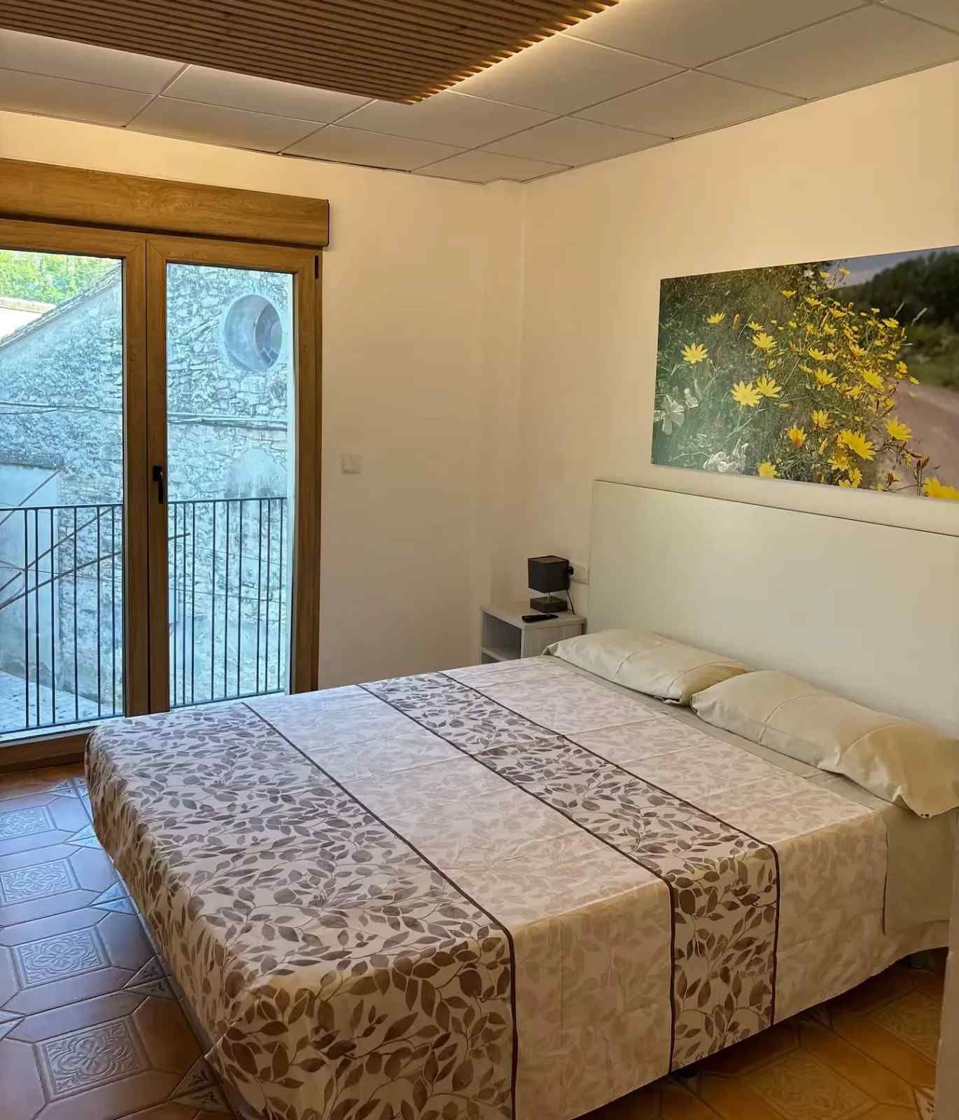 Dormitorio con cama doble, acceso al balcón, edredón con estampado floral, obras de arte y lámpara de techo texturizada.