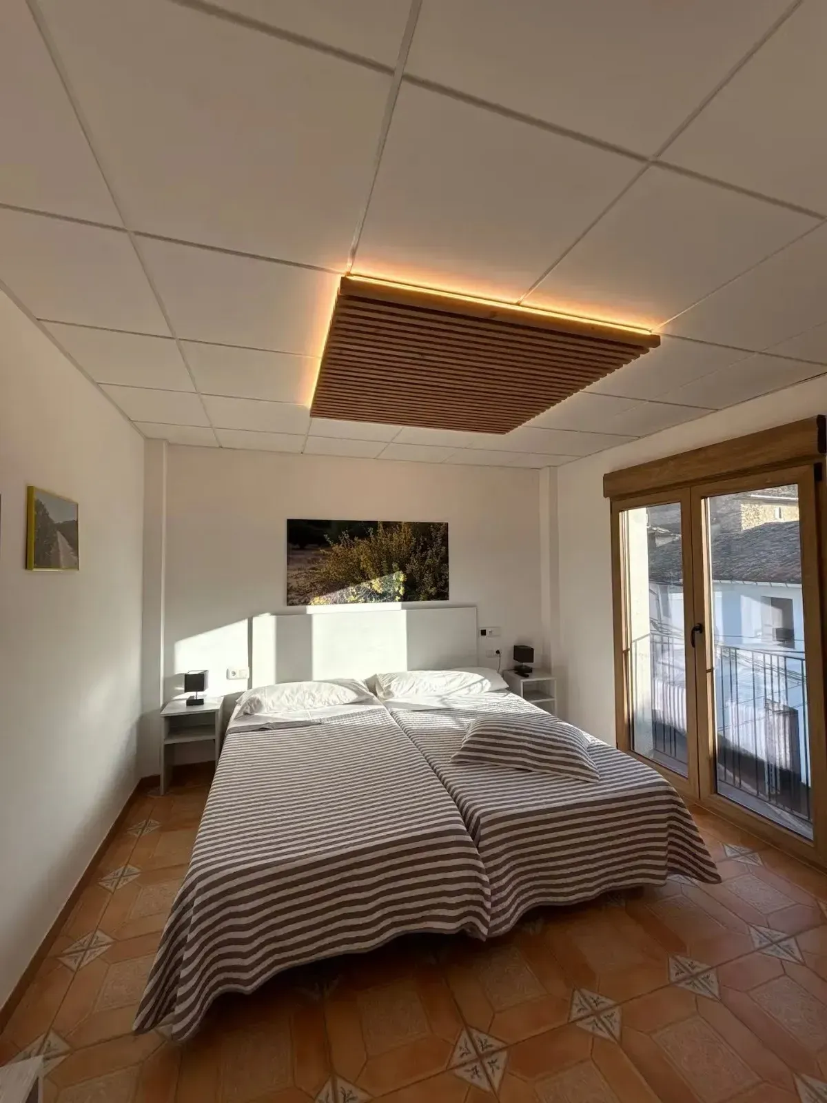 Dormitorio con cama estampada, luz de techo, suelo de baldosas y puertas de balcón.