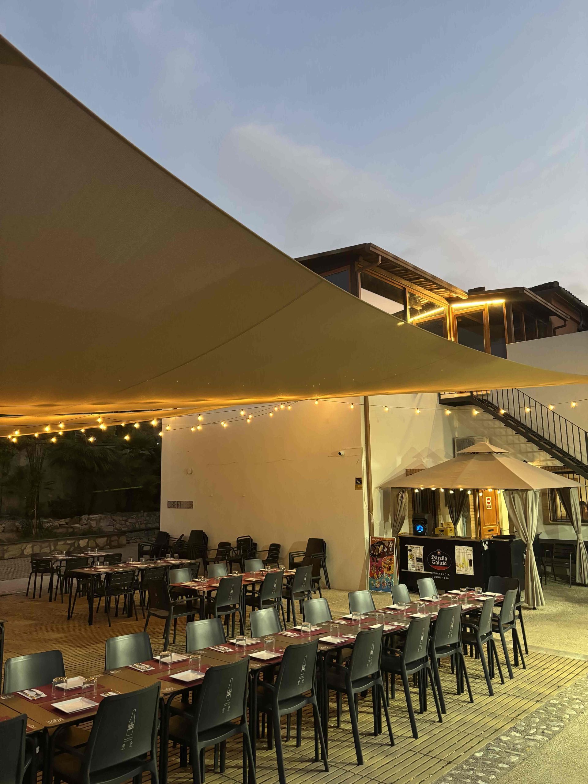 Zona de estar al aire libre cubierta con techo de madera, paredes de vidrio y muebles grises.