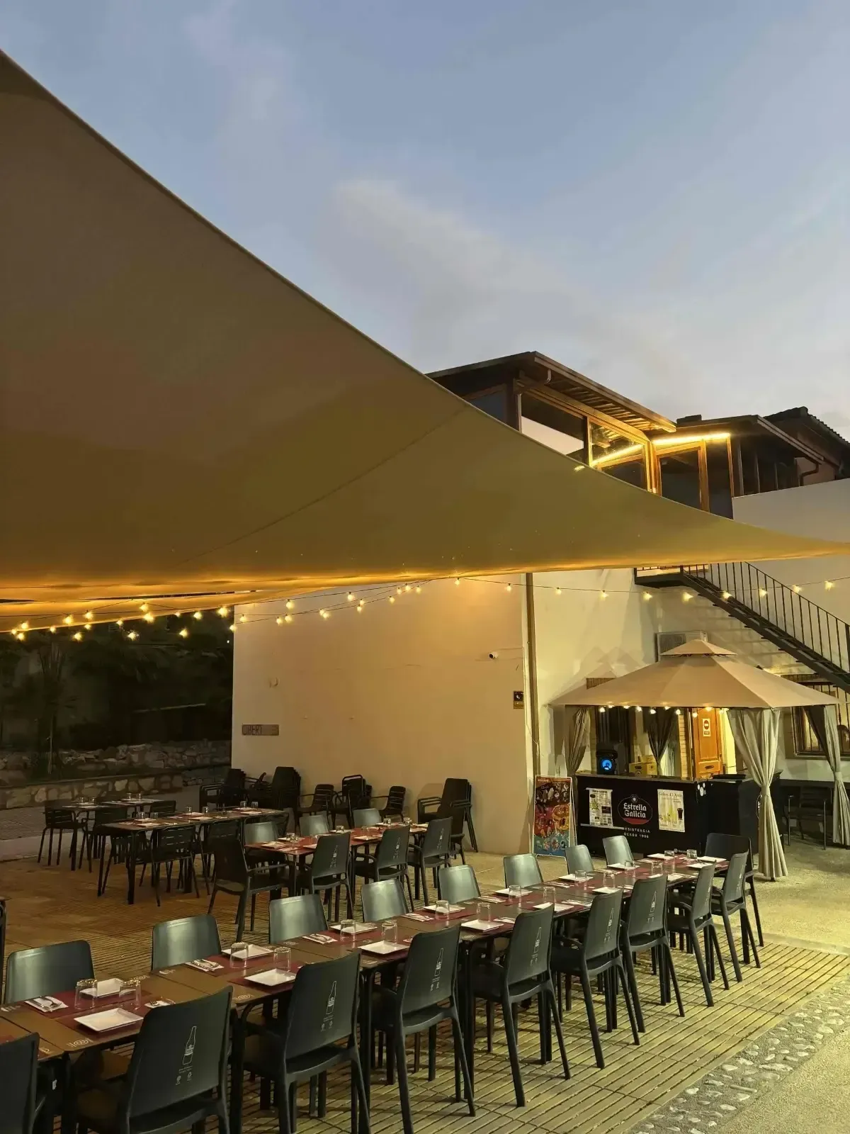 Zona de estar al aire libre cubierta con techo de madera, paredes de vidrio y muebles grises.