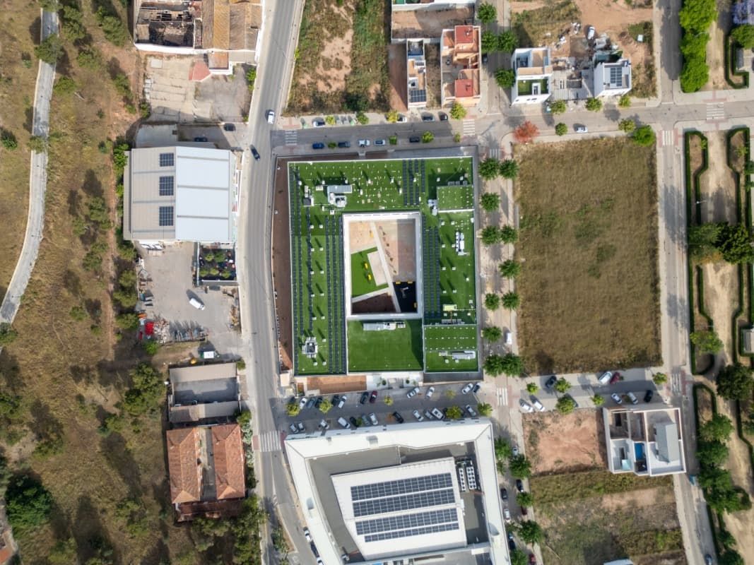 Vista aérea de un edificio moderno con techo verde y patio central, rodeado de carreteras, edificios y terrenos no urbanizados.