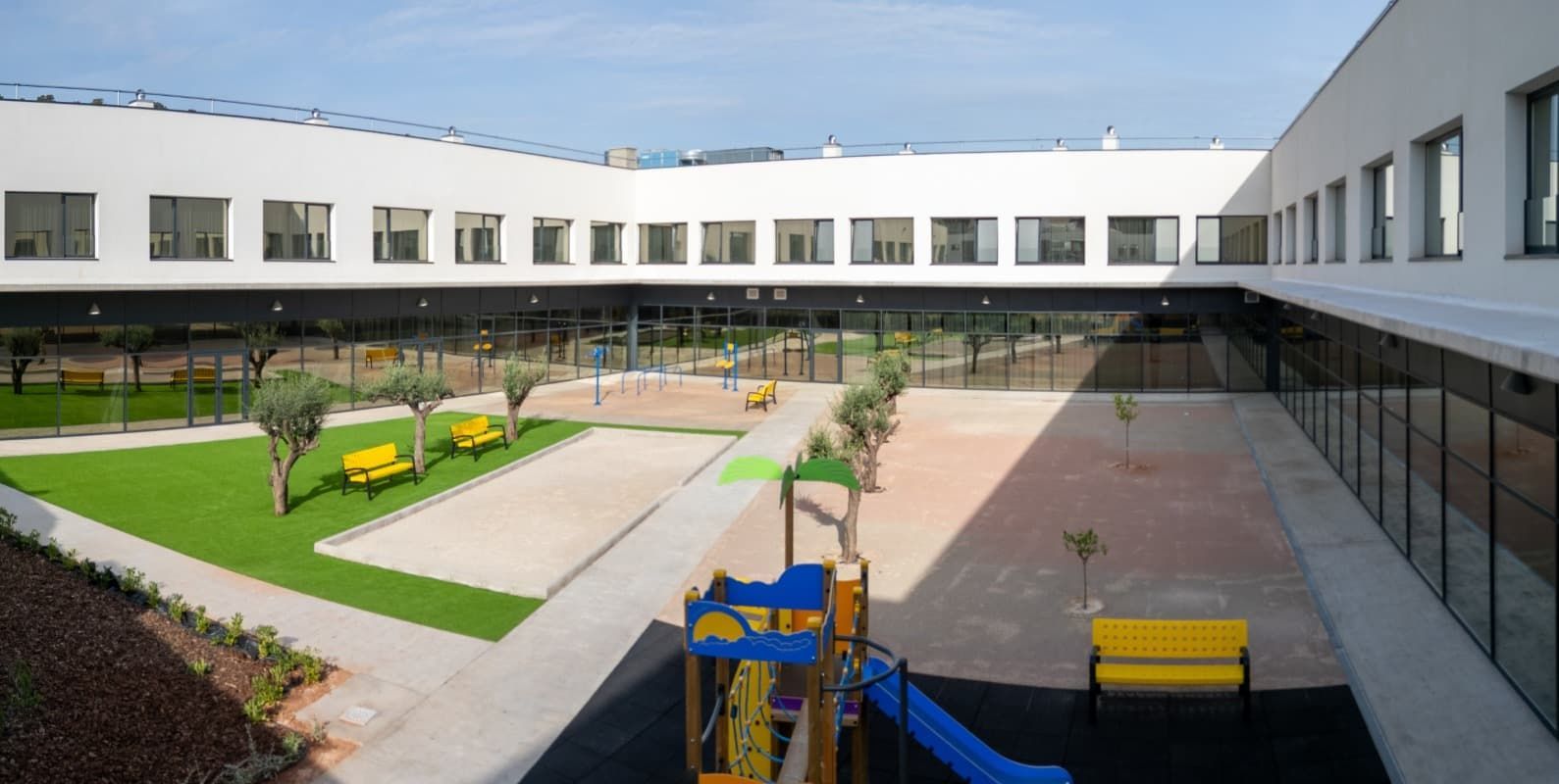 Patio exterior de edificio blanco, con zona de juegos y césped verde.
