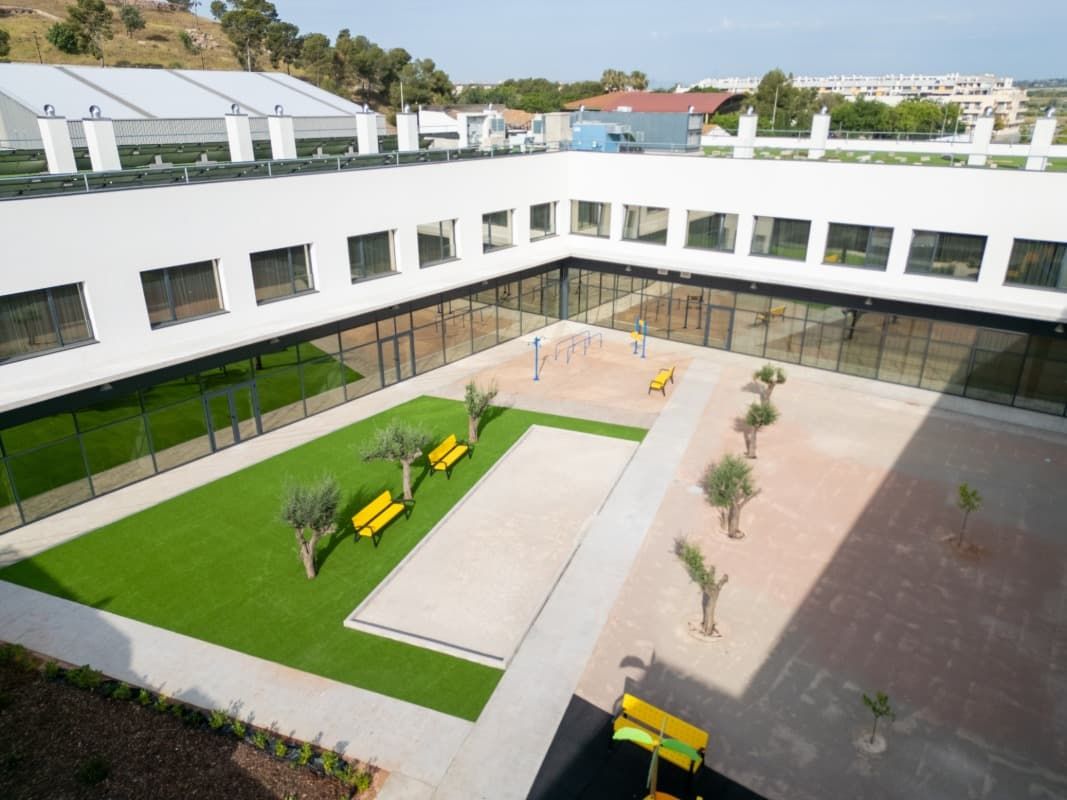 Patio con espacio verde, bancos, árboles y un edificio blanco y moderno que lo rodea.