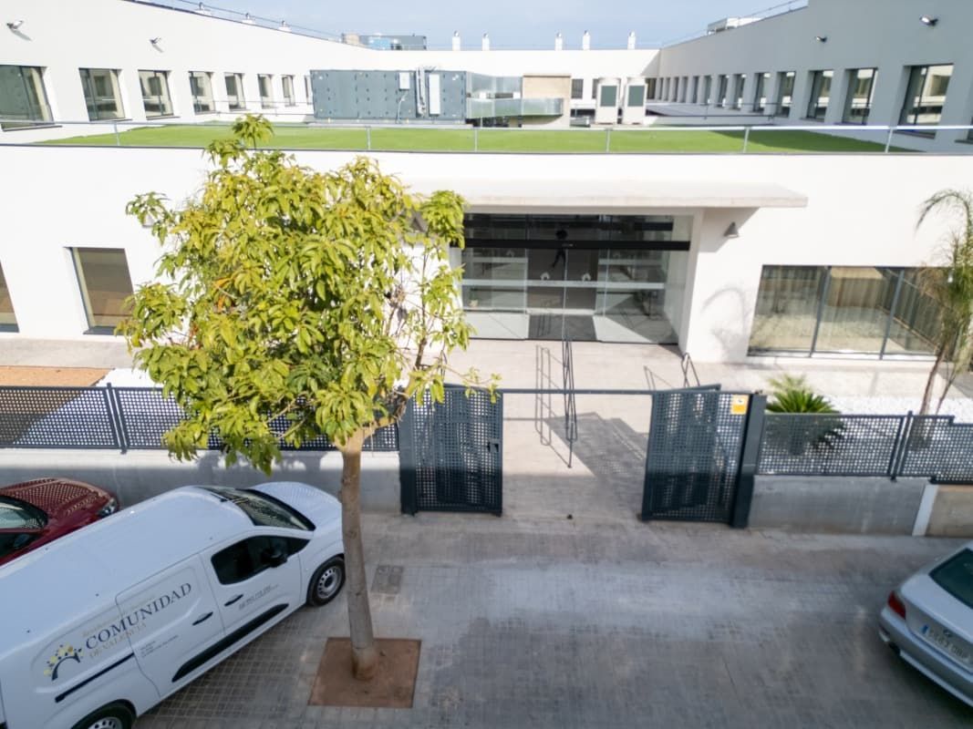 Entrada de edificio blanco con techo verde, árbol y camioneta de reparto en área de estacionamiento.