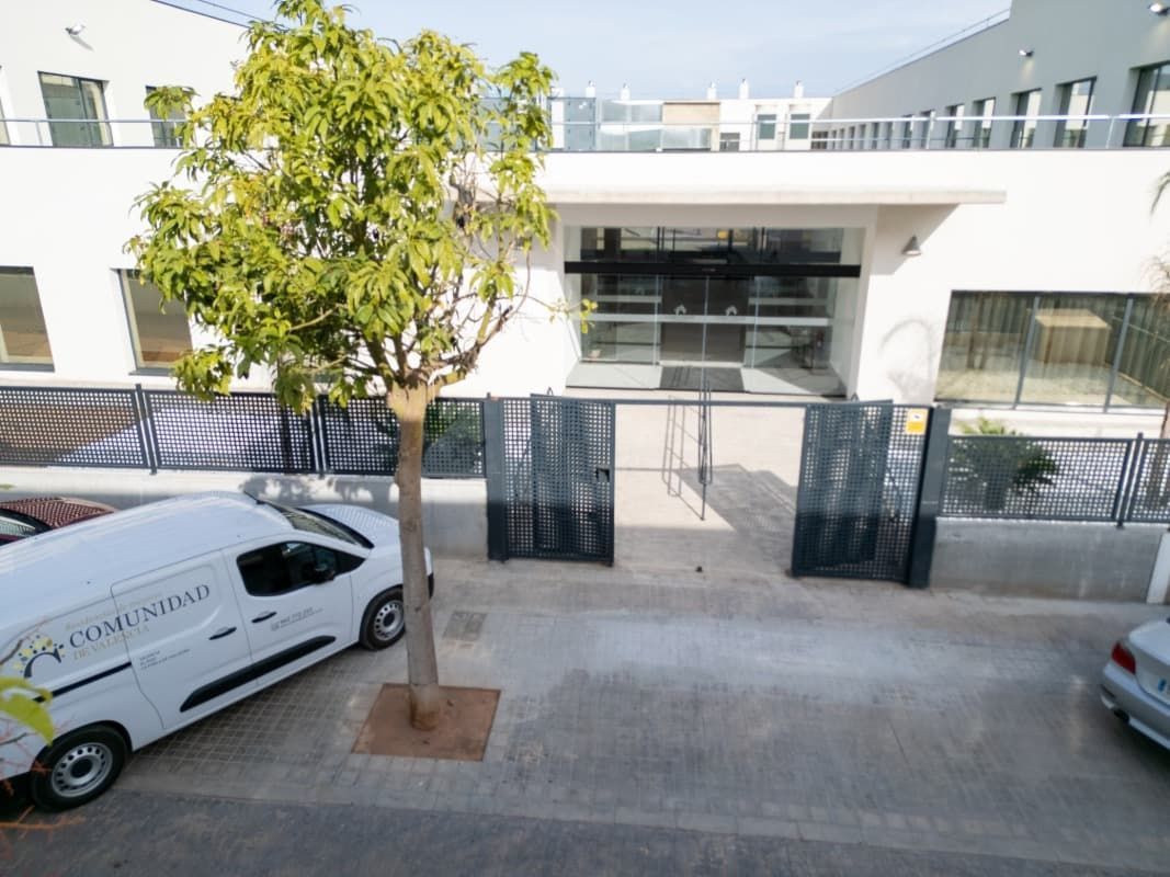 Entrada de edificio blanco con portones negros abiertos, un árbol pequeño y una camioneta blanca estacionada en la calle.