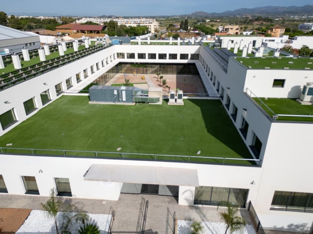 Vista aérea de un edificio blanco con techo verde, patio cuadrado en el centro y un paisaje urbano al fondo.