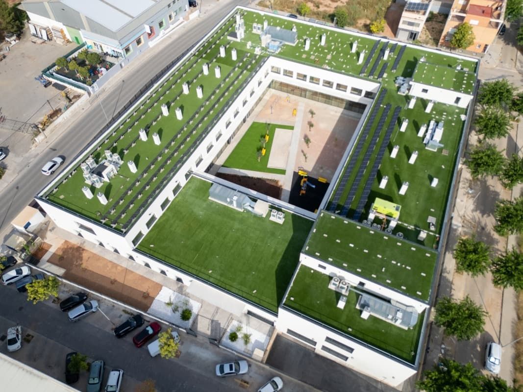 Vista aérea de un gran edificio blanco con techo verde y patio central. Coches aparcados cerca.