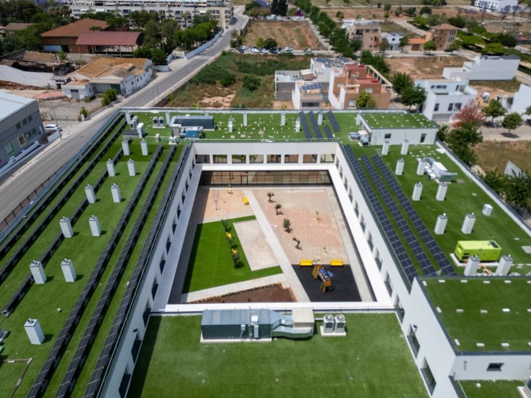 Vista aérea de un edificio blanco con patio central, techos verdes y paneles solares en una zona residencial.