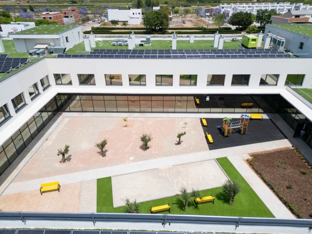 Vista aérea de una escuela moderna con patio abierto, techo verde, paneles solares y zona de juegos.