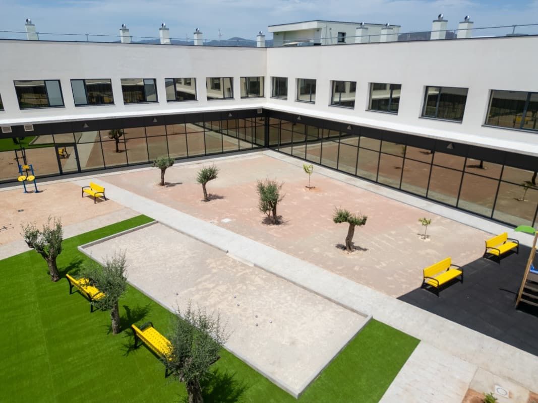 Patio con árboles, bancos amarillos y zona de juegos. Edificio blanco con paredes de cristal.