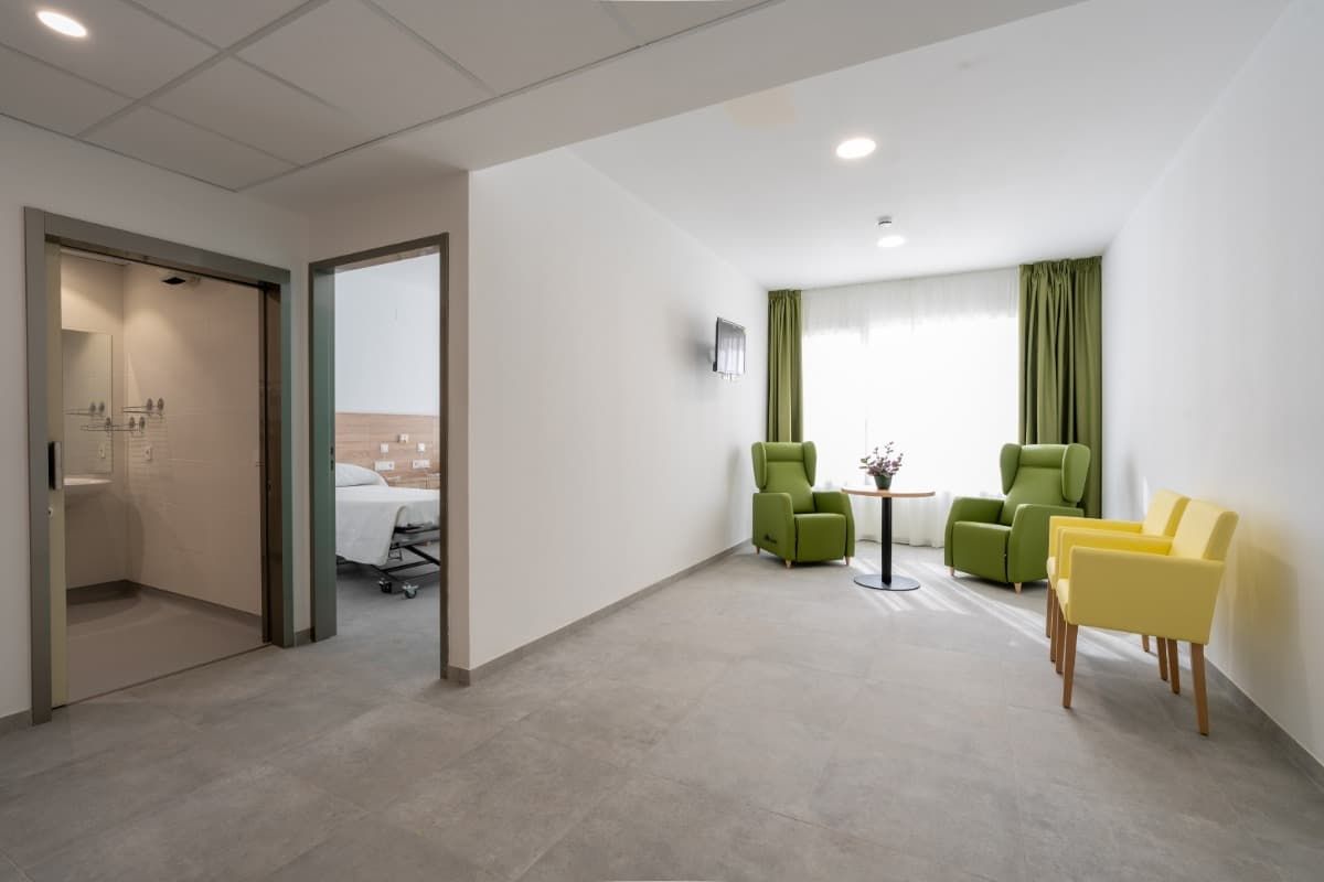 Sala de espera con sillones, una pequeña mesa y vista a una habitación de hospital.