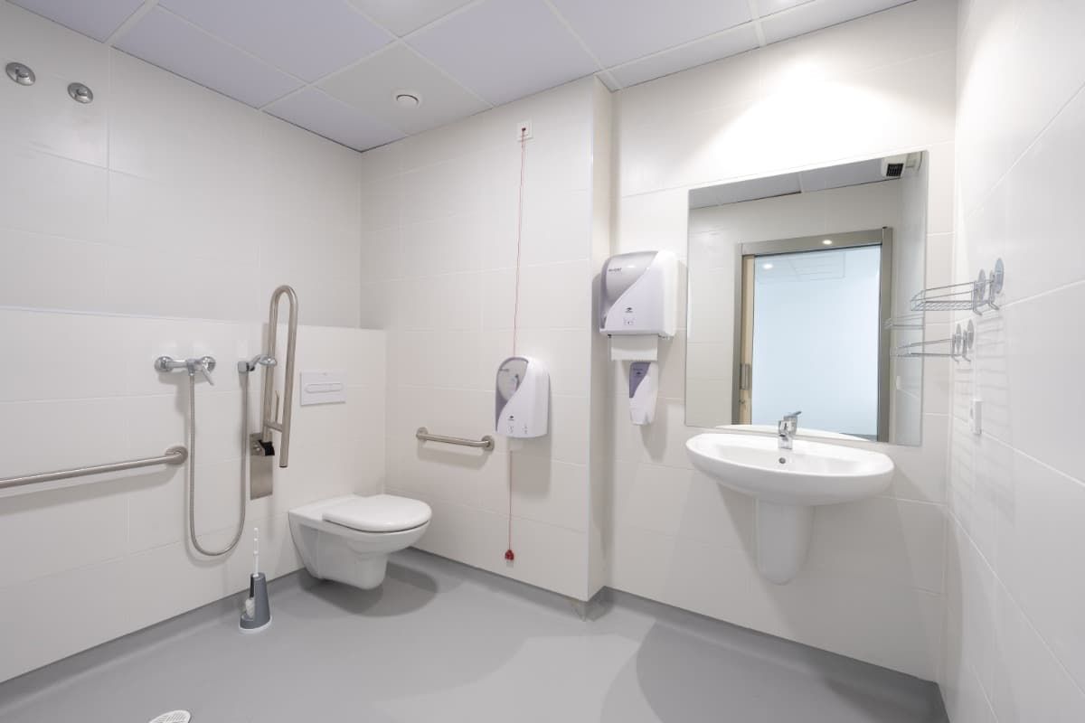 Baño público moderno y accesible. Paredes blancas, suelo gris, inodoro con barras de apoyo, lavabo y espejo.