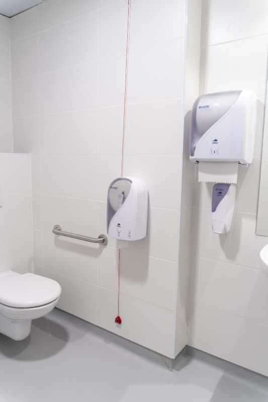 Un baño blanco con inodoro, dispensadores de toallas de papel y barra de apoyo. Un reguero de toallas de papel rojas cuelga.