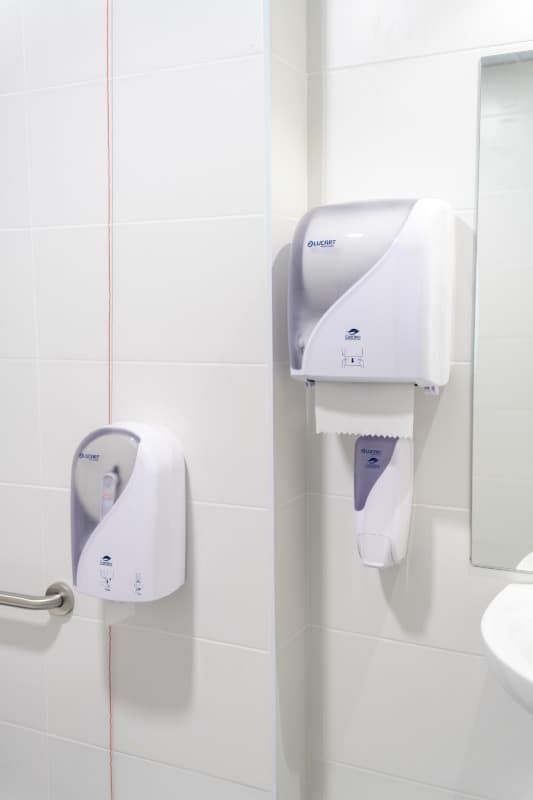 Baño blanco con dispensadores de toallas y papel higiénico en pared alicatada.