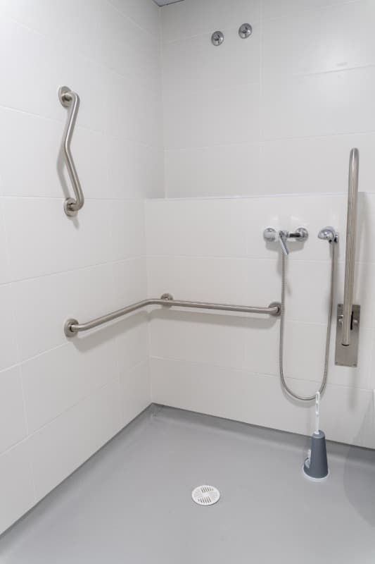 Ducha accesible con barras de apoyo y cabezal de ducha de mano, piso gris, paredes blancas.