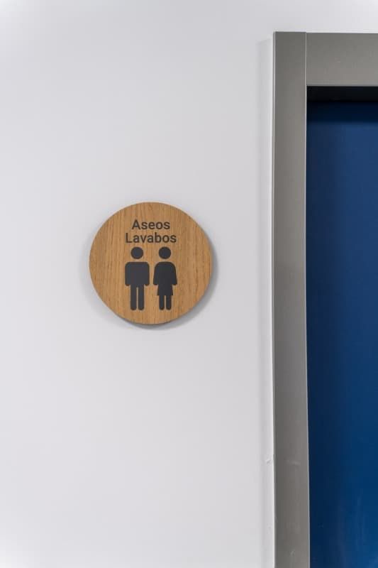 Letrero de baño de madera con figuras masculinas y femeninas, en una pared blanca al lado de una puerta azul.