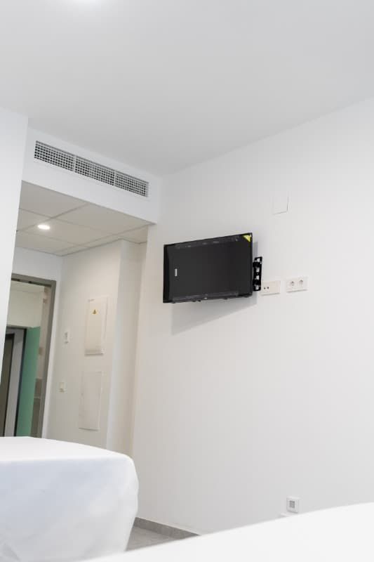 Televisor montado en una pared blanca, encima de una cama. Enchufes junto al televisor.