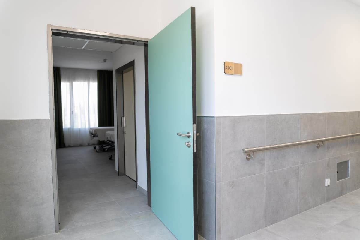 Puerta verde azulada abierta que da a una habitación. Vista interior: pasillo con pasamanos, habitación con ventana.