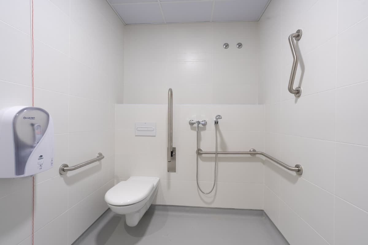 Baño accesible con inodoro, barras de apoyo y ducha. Paredes blancas, suelo gris y grifería de acero inoxidable.
